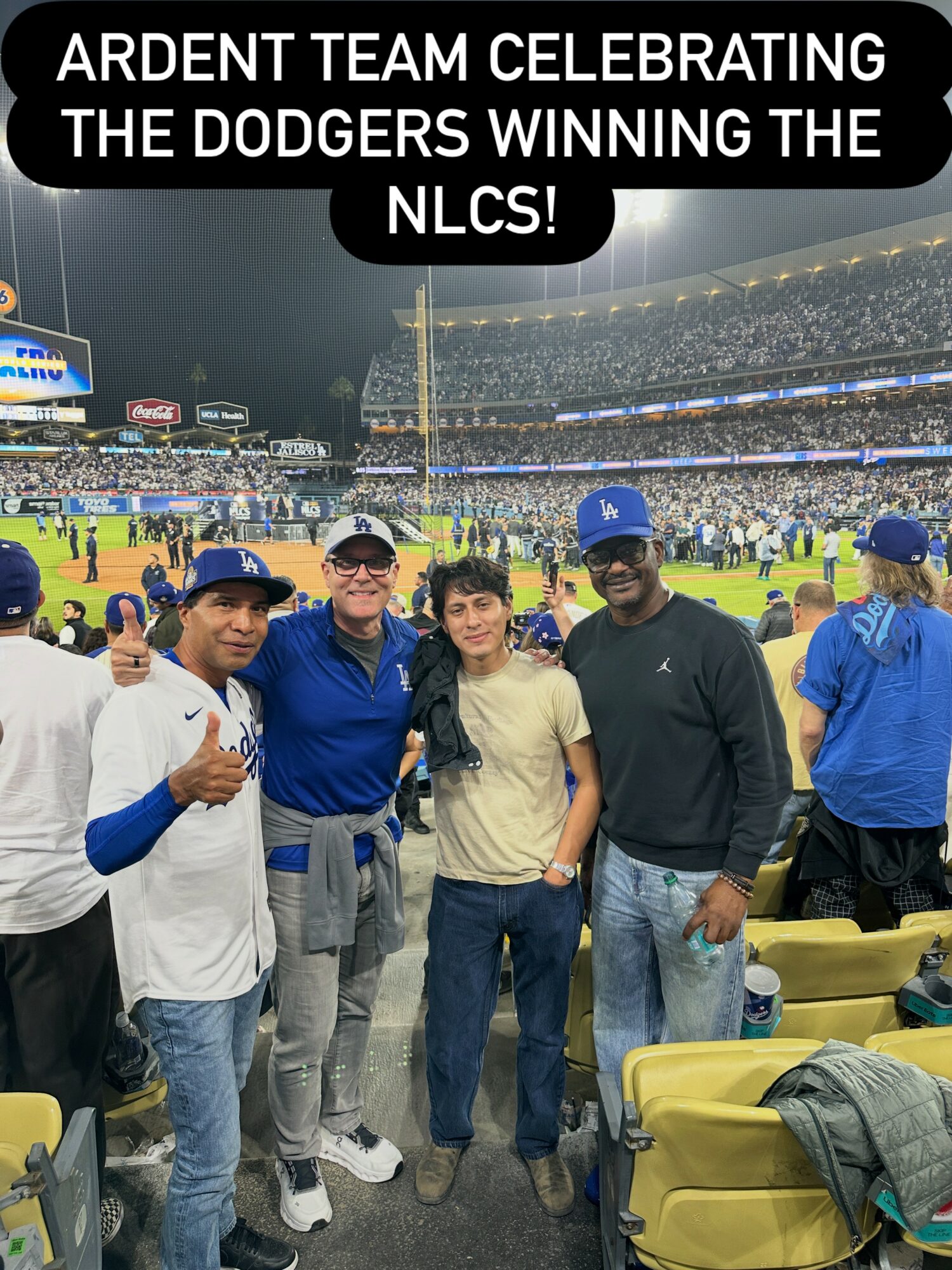 Four people standing together at a stadium, celebrating a baseball victory, with a crowd and field in the background.