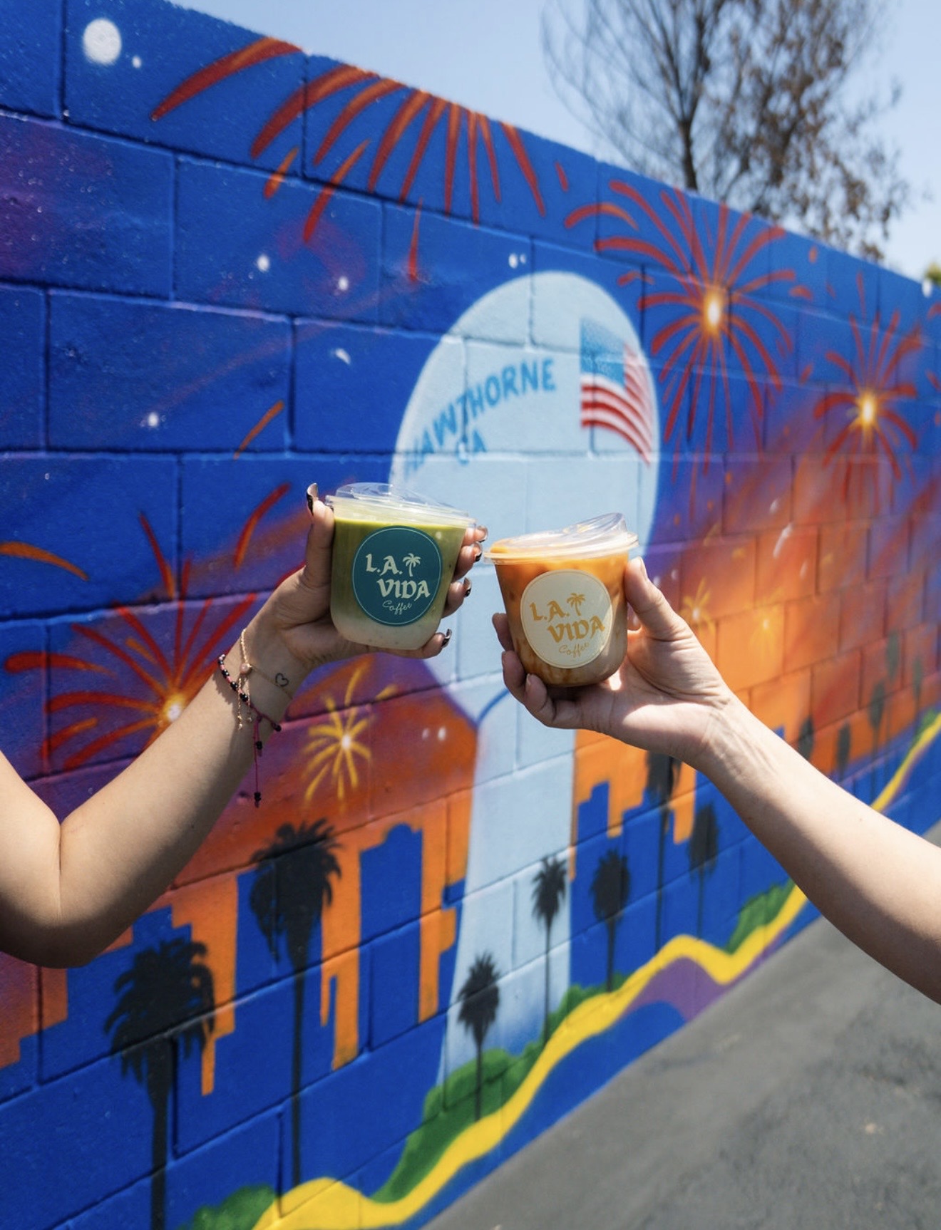 Two people hold cups in front of a colorful mural with fireworks and a city skyline, blue sky background.