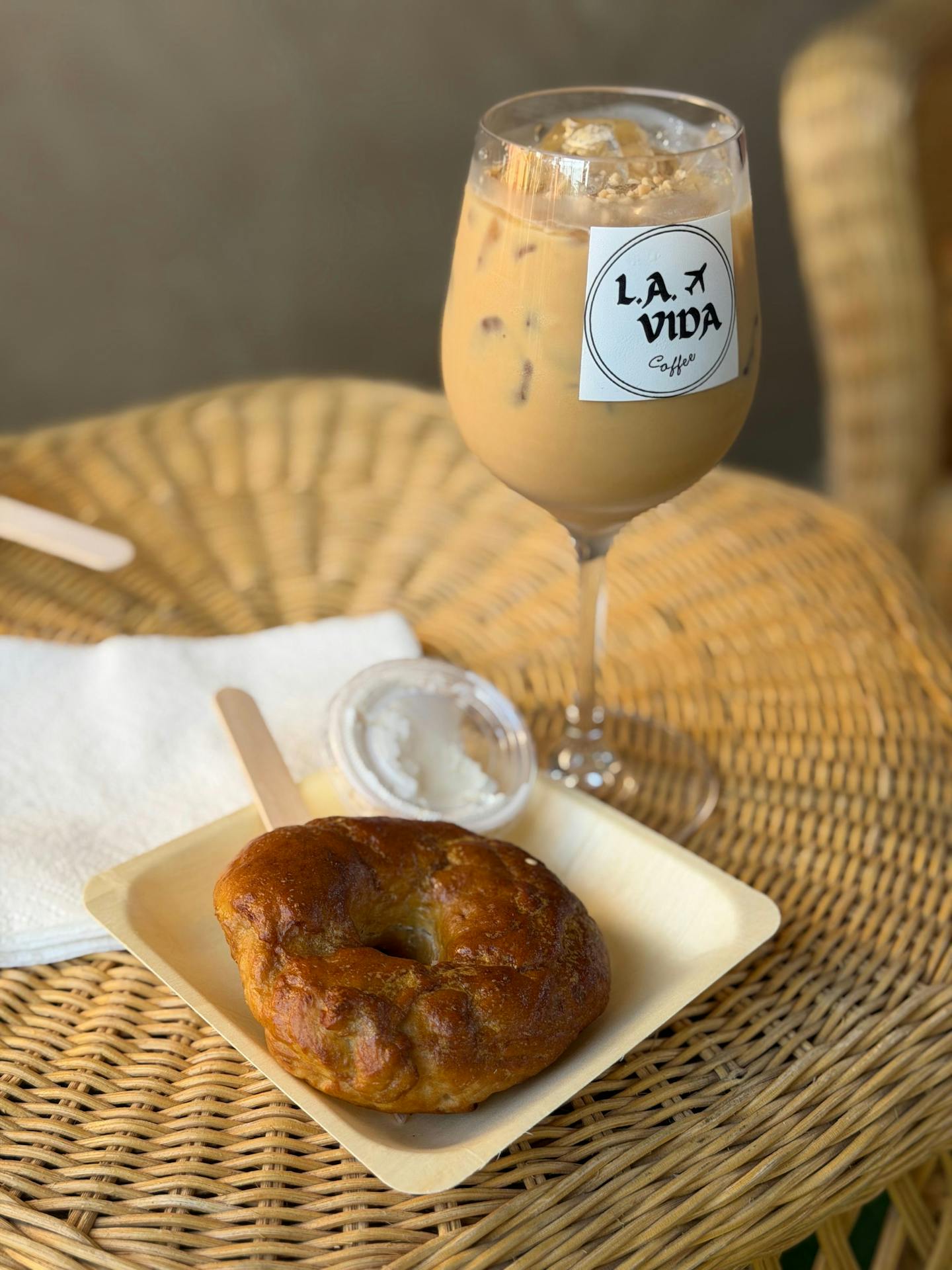 Glass of iced drink with a label, and a fried pastry on a white plate on a wicker table.