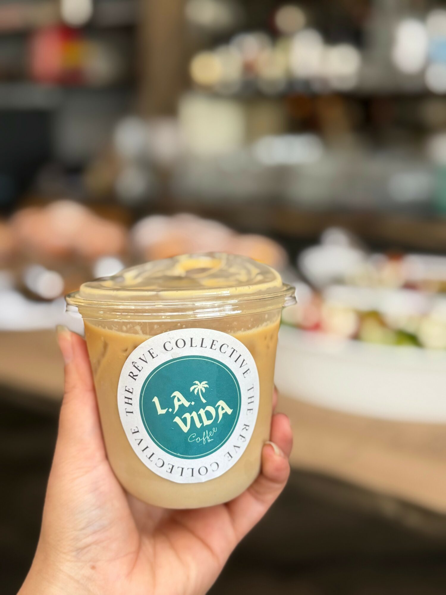 Hand holding a cup of dessert with a label reading 'L.A. Vida' in a bakery or cafe setting.