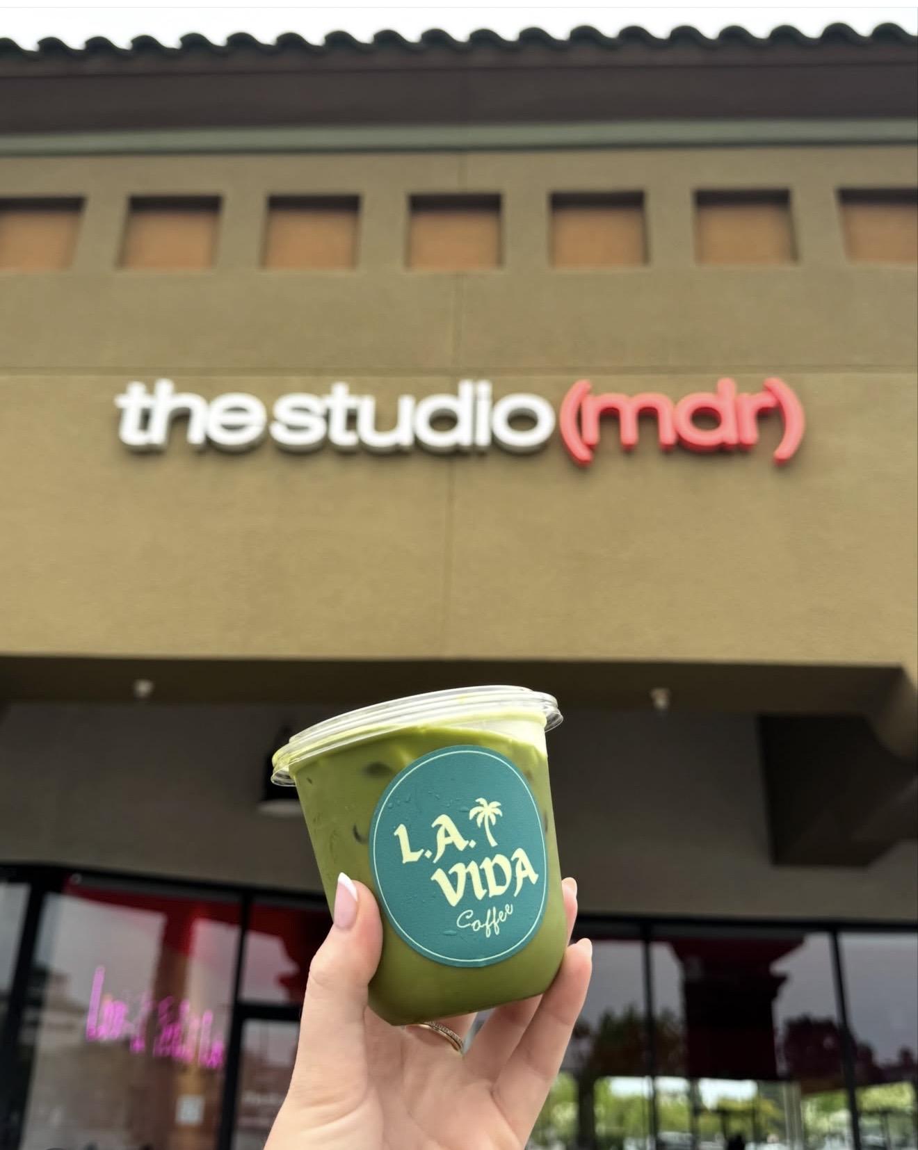 Hand holding a green smoothie cup with a blue logo in front of a building with a sign reading 'the studio (mdr)'.