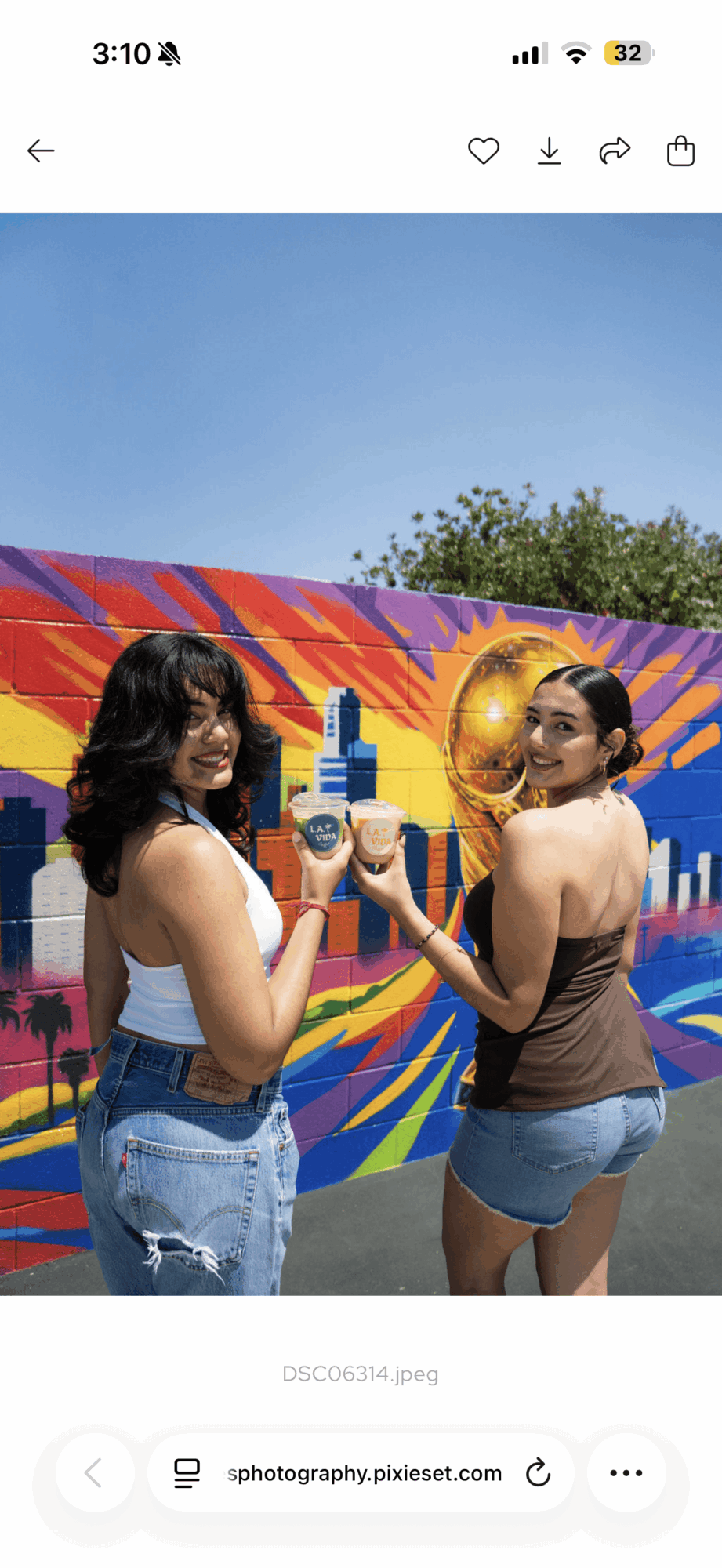 Two women stand in front of a colorful mural, holding a small object, with trees and sky in the background.