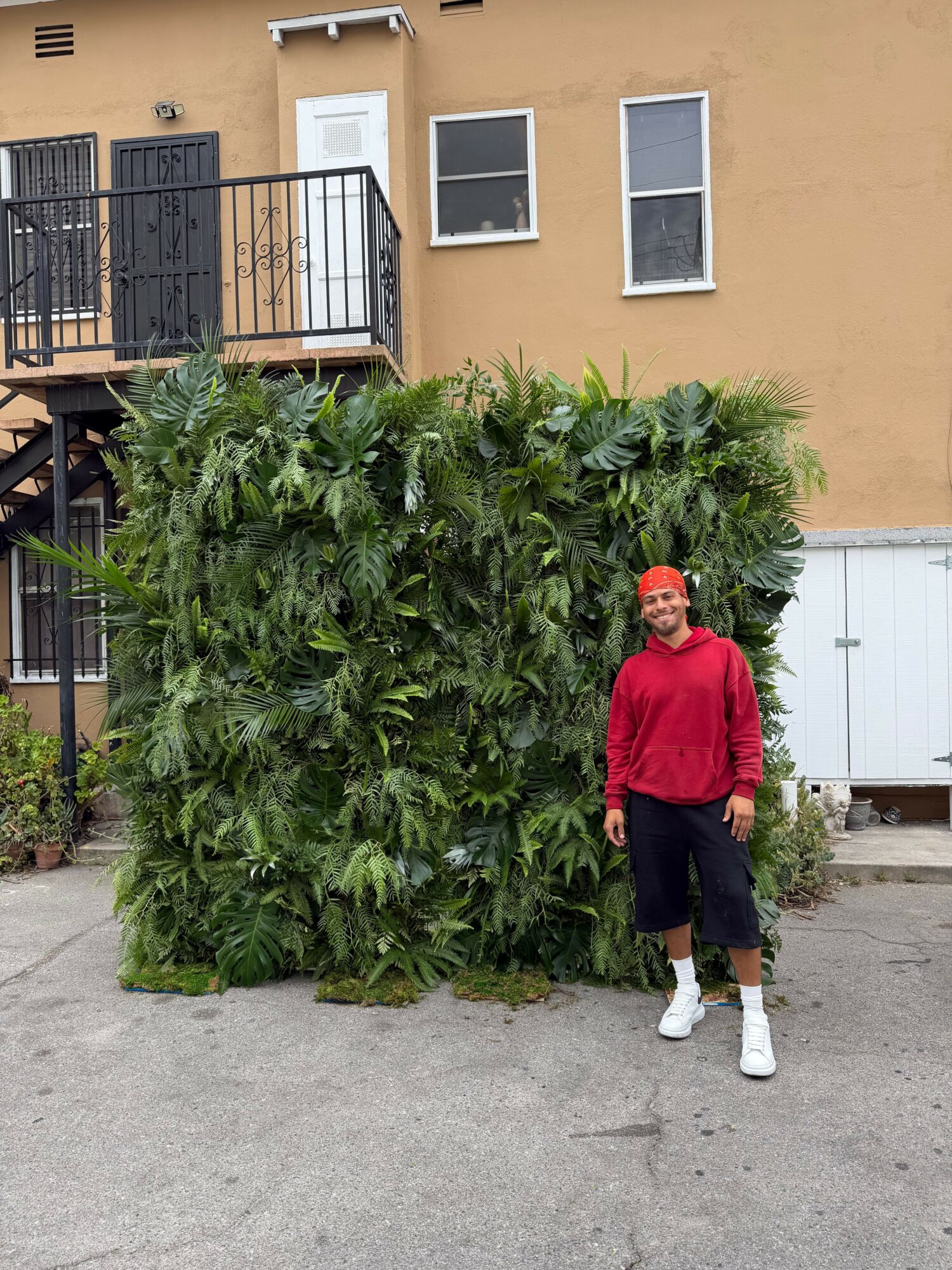 Person in red jacket and black pants standing next to large green bush outside building.