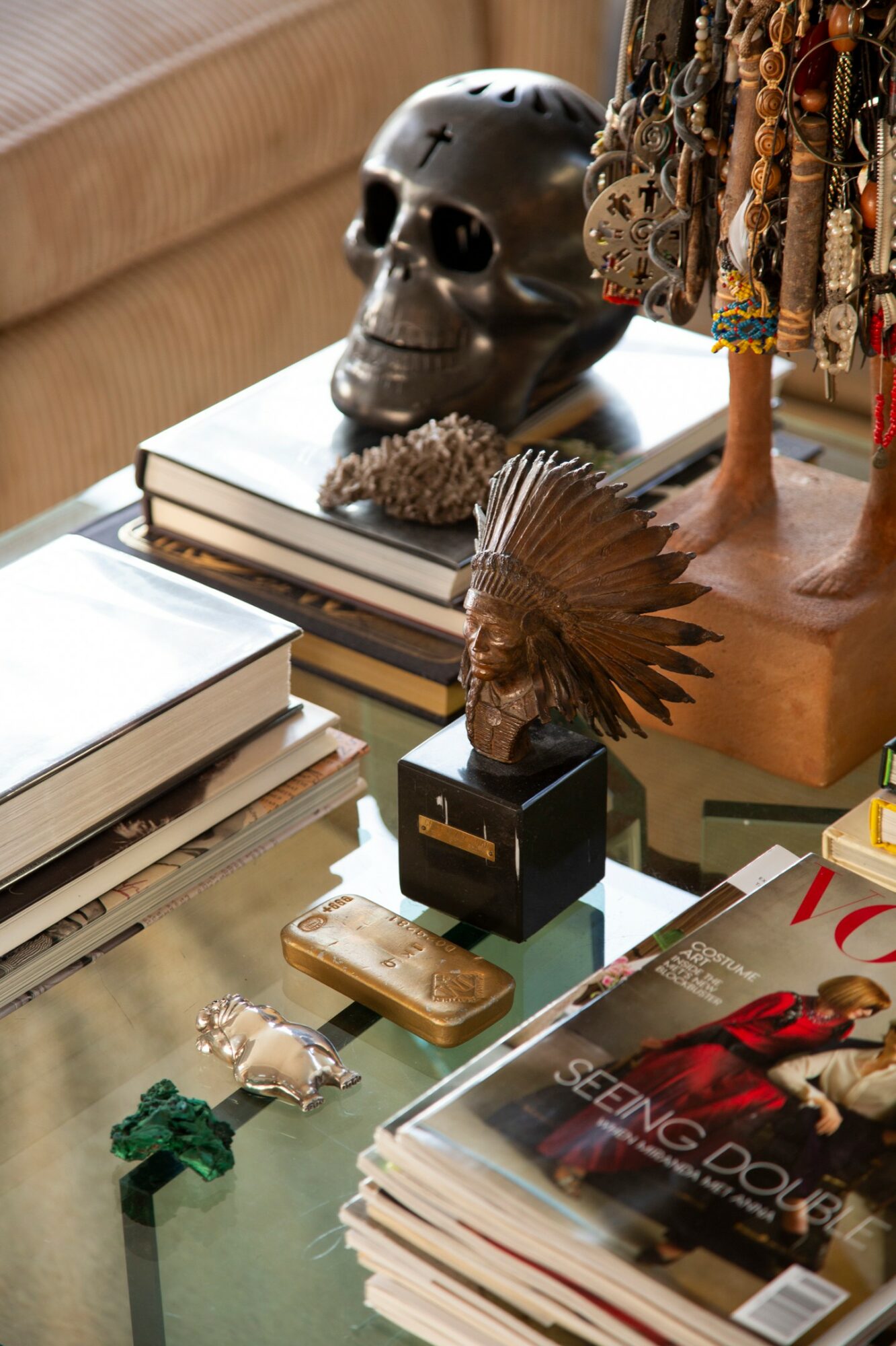 Decorative skull, jewelry, and books on a glass table with a magazine, small figurines, and a wooden stand.