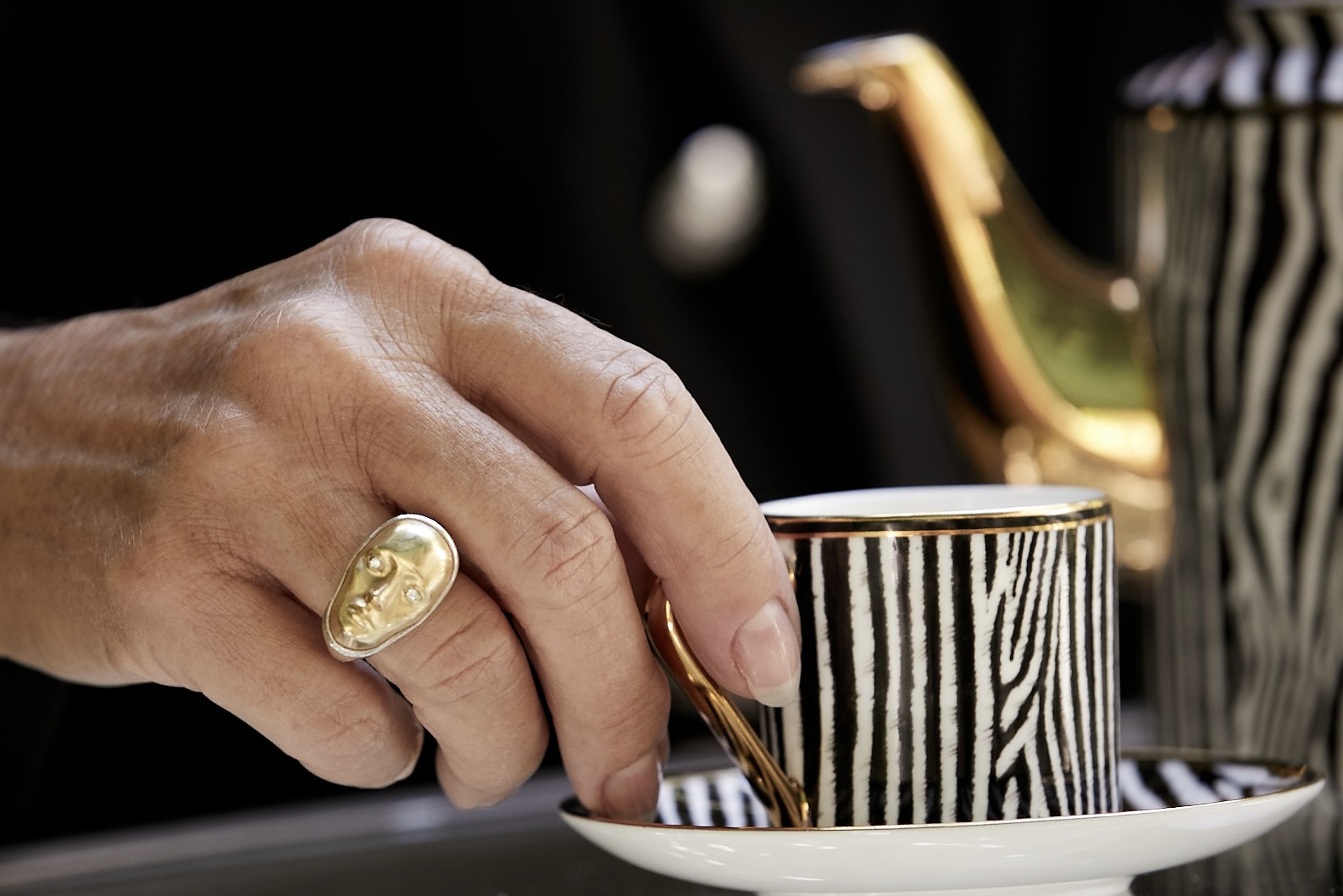 Hand with a ring holding a gold utensil over a black and white striped cup and saucer, with a teapot in the background.