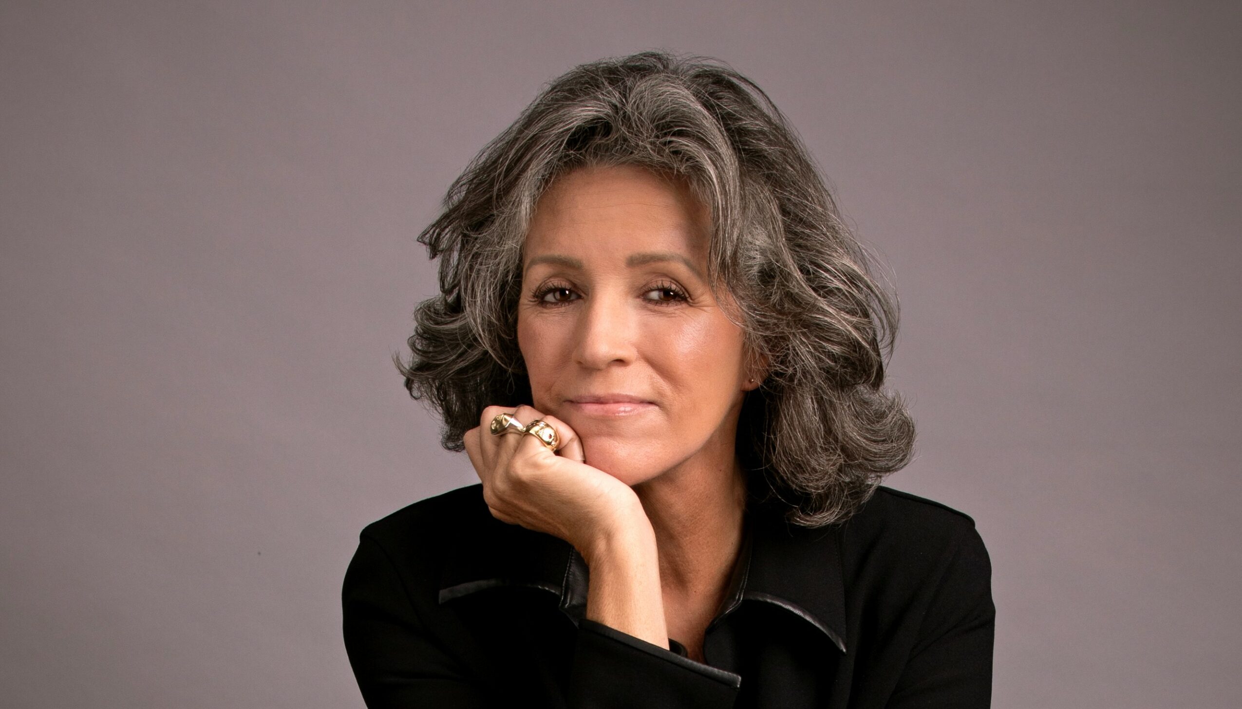 Portrait of a woman with gray hair resting her chin on her hand, wearing a black top, against a plain background.