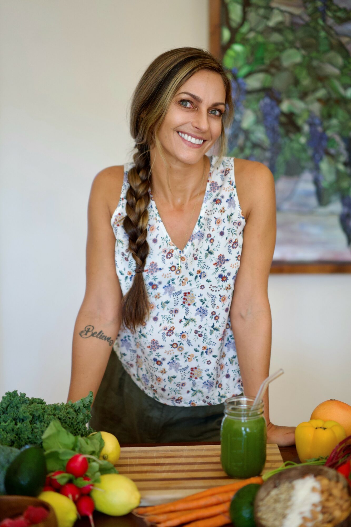Smiling woman with long braided hair standing behind a table with vegetables and a green smoothie, in a room with a painting.